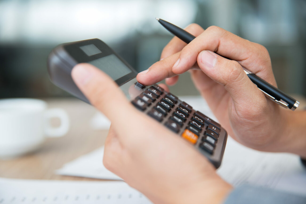 Imagem aproximada mostra mão masculina segurando e digitando em calculadora