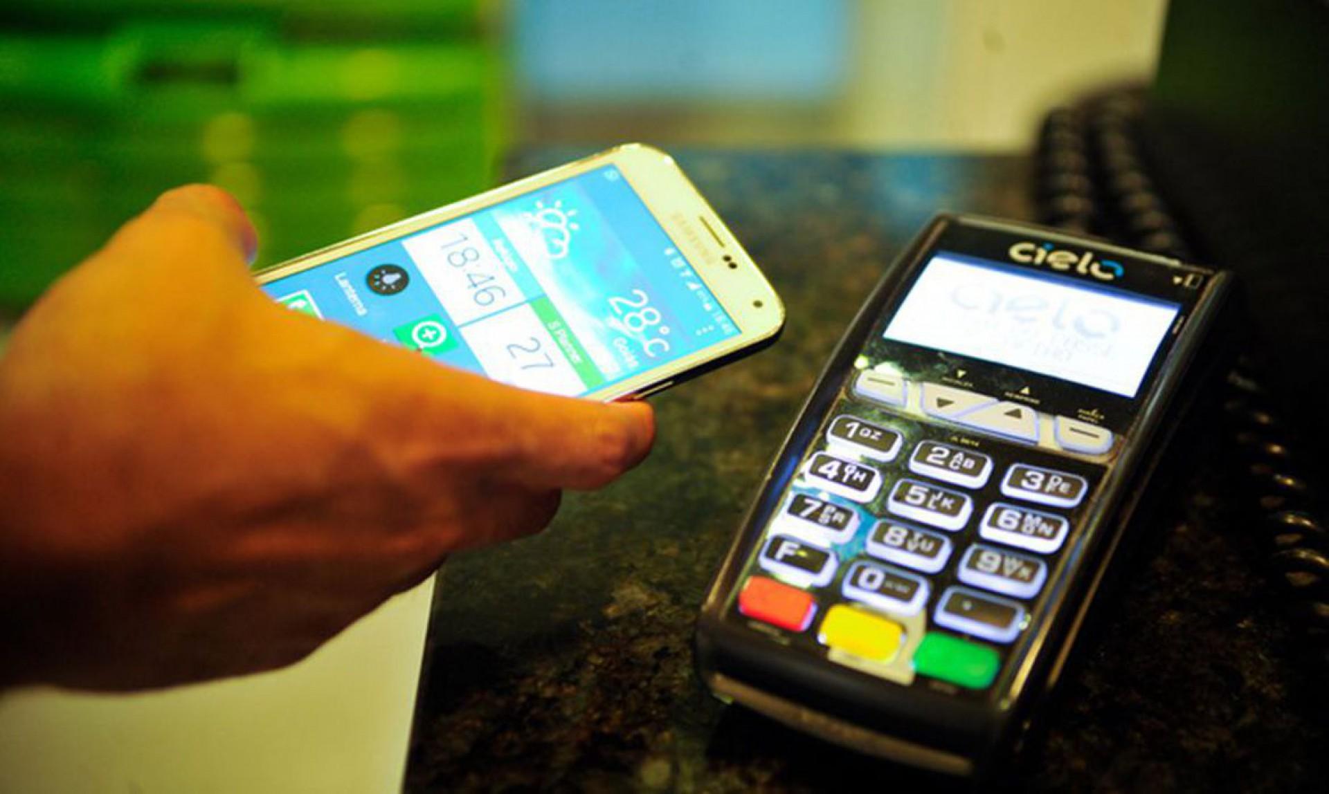 Foto mostra homem segurando celular próximo de máquina de cartão de crédito.