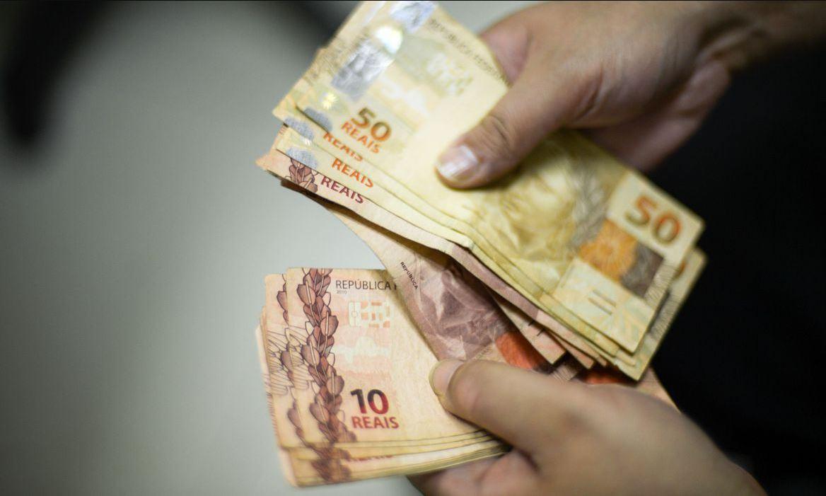 Foto com mãos de homem segurando e contando diversas notas de 50 reais e 10 reais