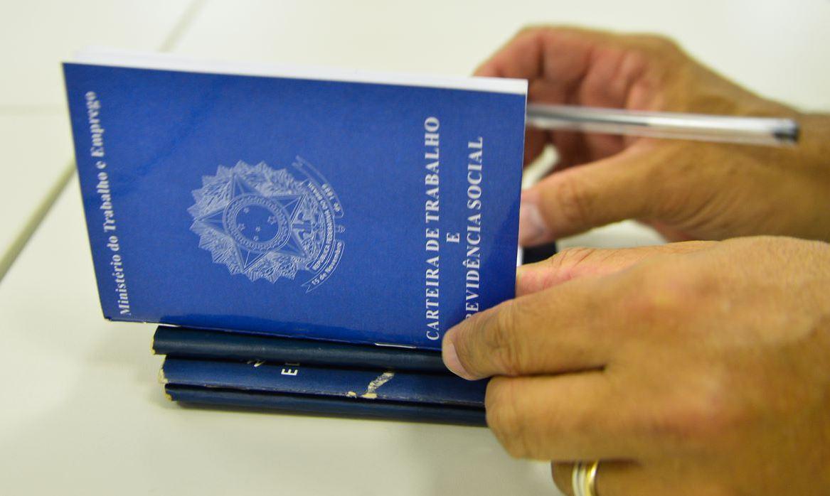 Foto mostra mãos de homem assinando carteiras de trabalho sobre uma mesa branca