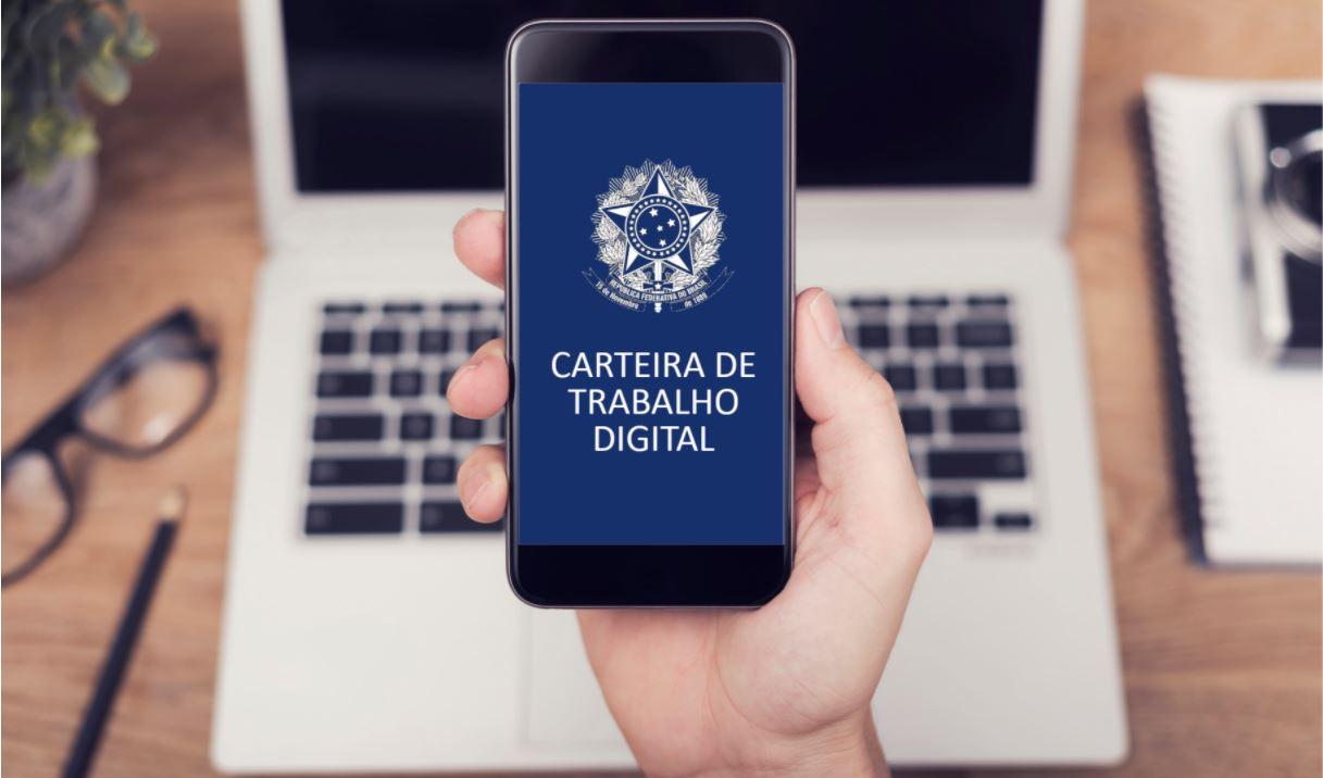 Foto mostra mãos segurando celular com aplicativo da carteira de trabalho digital aberta, e um notebook cinza aberto sobre a mesa de madeiraao fundo
