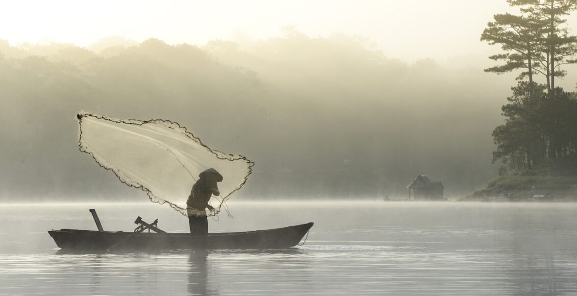 Foto mostra pescador sobre barco em meio ao mar atirando rede de pesca.