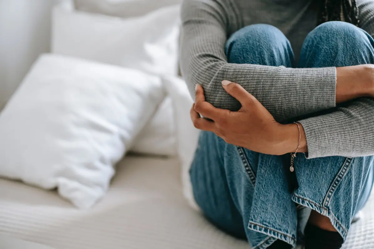 Imagem mostra pessoa de calça jeans sentada em uma cama, segurando as próprias pernas, como um sinal de dor provocado pela Intoxicação alimentar