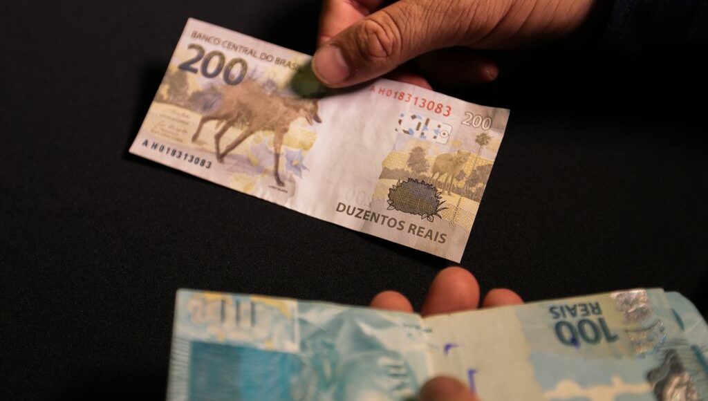 Foto mostra homem segurando notas de 200 e 100 reais.